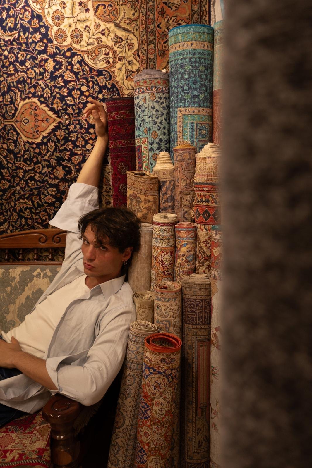 The artisan reclines on a carved settee beside towering rolled rugs